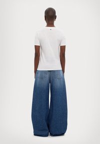 Valkoinen lyhythihainen t-paita, jossa on pieni logo takana, yhdistettynä ylisuurisiin sinisiin farkkulegginsseihin, joissa on kaksi takataskua.