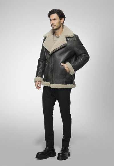 Giacca in shearling nera in pelle con colletto e polsini in pelliccia beige. Presenta chiusura con zip e tasche laterali. Il modello indossa pantaloni neri e stivali.