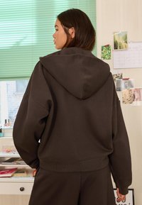 Smeđa majica s kapuljačom opuštenog kroja, izrađena od teksturiranog materijala, s rebrastim manšetama i rubom, prikazana s leđa na svijetloj pozadini.