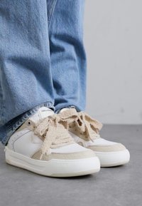 White sneakers with beige accents, textured materials, large laces, and a smooth finish. Worn with light blue denim jeans.