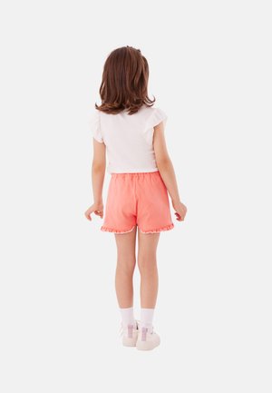 Meisje vanaf de achterkant gezien, gekleed in een witte top met ruches aan de mouwen, koraalroze shorts met ruches aan de zoom, witte sokken en witte sneakers tegen een effen achtergrond.