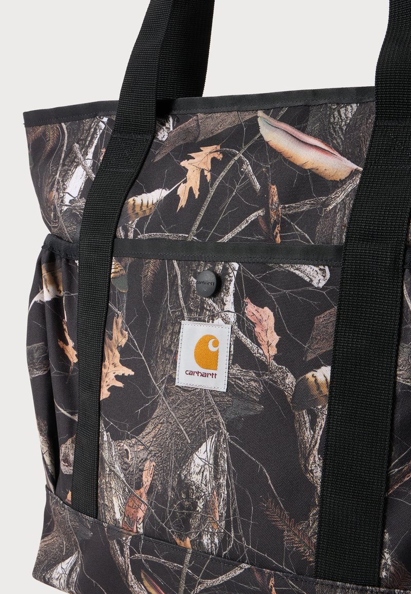 Tote bag with black straps and camouflage pattern featuring branches and leaves, front pocket with Carhartt logo patch and snap button.