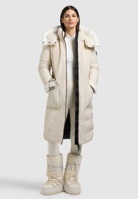 Langer cremefarbener Steppmantel mit Kapuze, die mit Lammfell gefüttert ist, schwarzem Reißverschluss, übergroßen Taschen und passenden hohen Schneestiefeln. Glattes, isoliertes Gewebe.