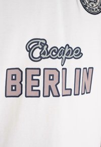 Țesătură albă cu cuvântul "Escape" scris cu litere cursive și "BERLIN" în litere mari tiparite în nuanțe de violet desaturat și contururi negre.