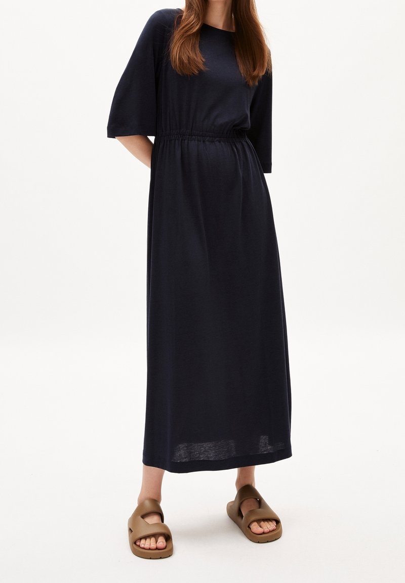 Laivastonsininen maksimekko, jossa on lyhyet hihat, nyörityksellä kiristetty vyötärö ja pehmeä kangas. Yhdistetty ruskeisiin liuku-sandaaleihin, joissa avoin varvas.