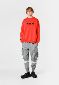 Sudadera roja brillante con letras negras "MWM", pantalones cargo grises con múltiples bolsillos y zapatos negros texturizados de tipo slip-on.