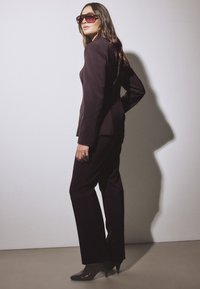 Woman in dark tailored suit and heeled boots poses sideways wearing large square sunglasses against a plain wall background.