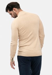 Beige Langarm-Strickpullover mit Rundhalsausschnitt, gerippten Bündchen und glatter Textur. In Kombination mit marineblauen Hosen, von hinten betrachtet.