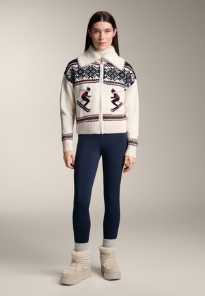 Krém színű gyapjú cipzáros pulóver műszőrme gallérral, síelés témájú és mintás kialakítással. Tengerészkék leggingsszel és bézs, texturált csizmával párosítva.