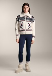 Beiger Wollpullover mit Reißverschluss und Pelzkragen, verziert mit Skimotiven und gemustertem Design. Kombiniert mit marineblauen Leggings und beigen, strukturierten Stiefeln.