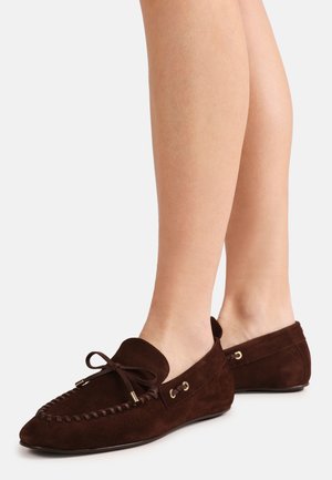 Mocassins en daim marron avec des accents de nœud portés pieds nus, vus de profil et de dos sur fond blanc.