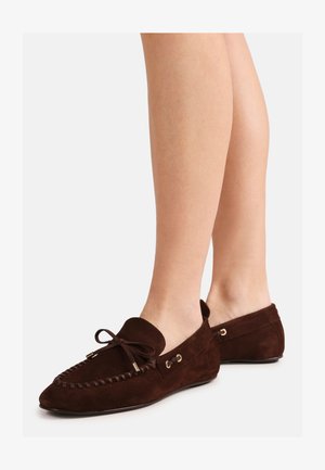 Brune loafers i semsket skinn med sløyfedetaljer, vist på bare føtter fra side- og bakvinkel på hvit bakgrunn.
