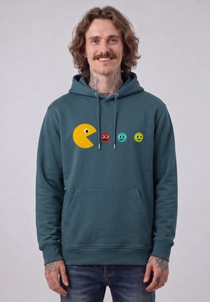 Mann mit Tattoos und Septumring trägt blauen Hoodie mit Pac-Man- und bunten Geistgesichtern, lächelnd vor einfachem Hintergrund.