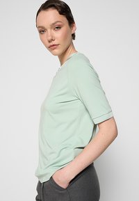 Lys grøn kortærmet t-shirt med rund hals, glat tekstur, diskret logo detaljer på siden og en let afslappet pasform.