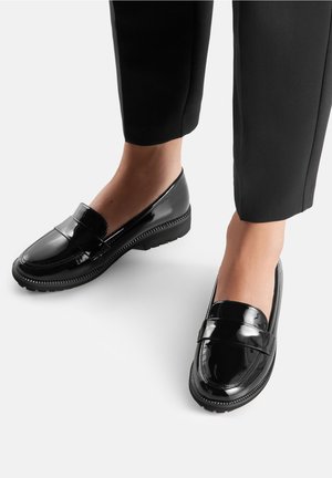 Person wearing black cropped pants and shiny black slip-on loafers with ridged soles on a light background.