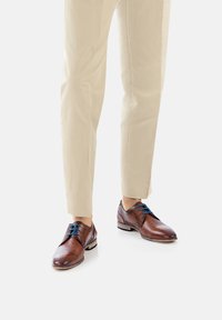 Chaussures habillées en cuir marron avec des lacets bleus, présentant un bout rond et une semelle noire contrastante, portées avec un pantalon beige clair.
