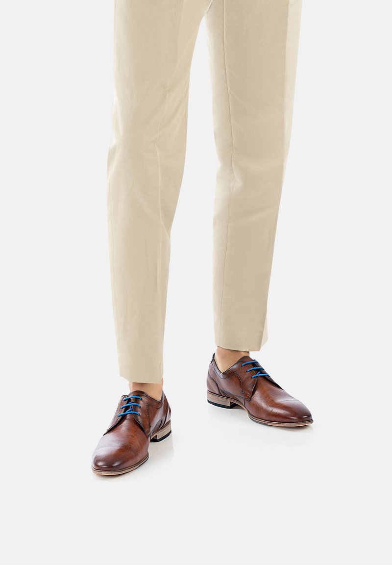 Chaussures habillées en cuir marron avec des lacets bleus, présentant un bout rond et une semelle noire contrastante, portées avec un pantalon beige clair.