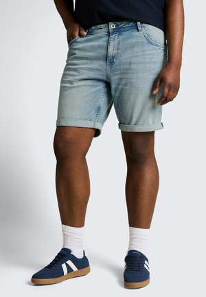 Persoon die lichtblauwe spijkershorts, witte sportsokken en donkerblauwe sneakers met witte strepen draagt, staand met één hand in de zak.