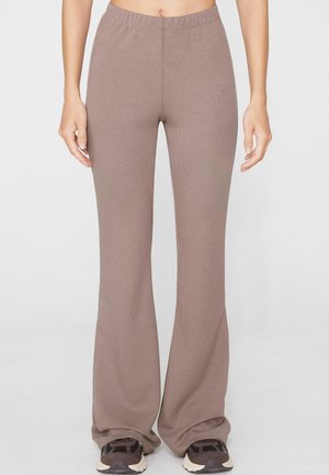 Persona che indossa pantaloni a coste color taupe a vita alta con gambe ampie che coprono leggermente scarpe da ginnastica scure, mostrata dalla vita ai piedi.