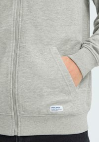 Sweat à capuche gris zippé avec poignets et ourlet côtelés, comportant deux poches avant et une étiquette en bas à gauche. Tissu doux et texturé.