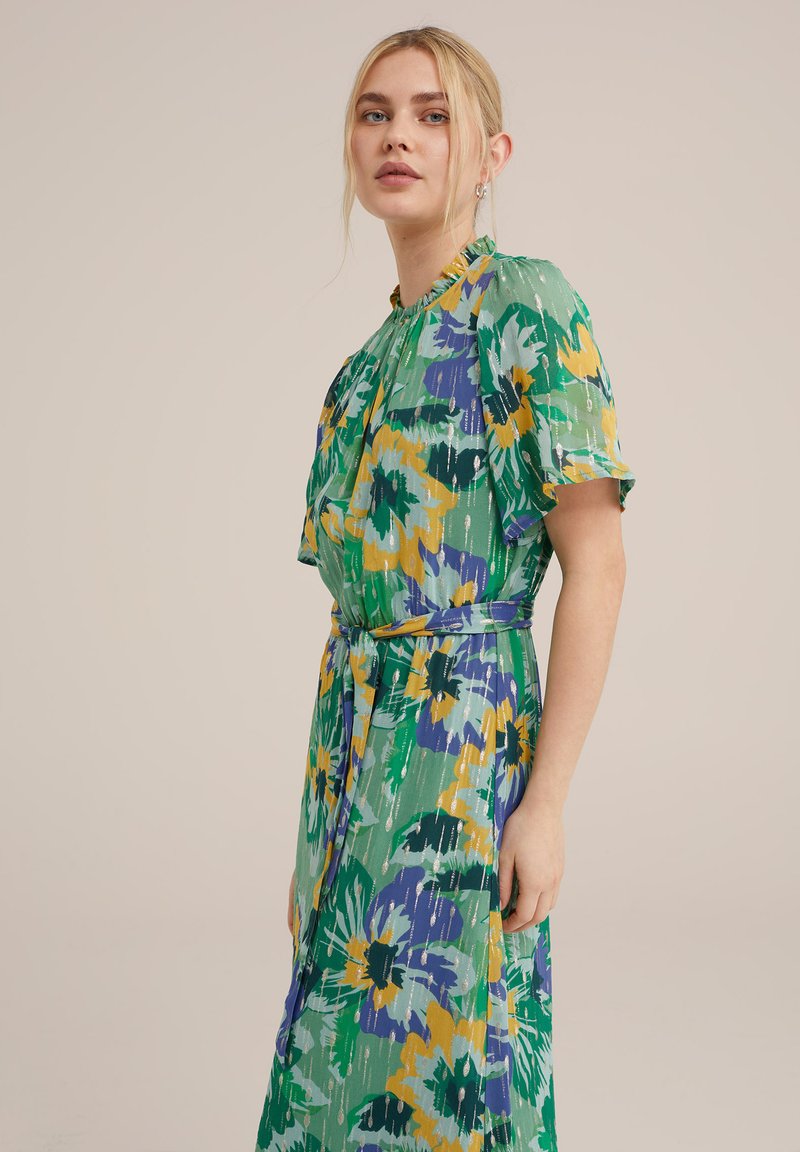 Blumenkleid mit grünem Grundton, verziert mit gelben und lila Blumen. Rüschenausschnitt und kurze Ärmel, Taillierung mit gebundenem Gürtel für eine schmeichelhafte Silhouette.