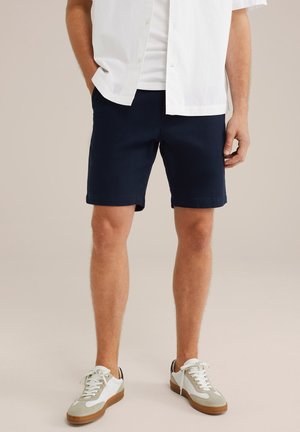 Shorts din bumbac de culoare bleu marin, cu o croială dreaptă, buzunare laterale și manșete răsucite. Asortate cu adidași albi cu accente gri.