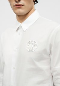 Άνδρας που φοράει λευκό πουκάμισο με κουμπιά και στρογγυλή ετικέτα λογότυπου Versace Jeans Couture στο αριστερό στήθος.