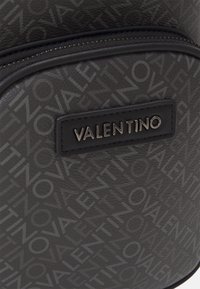Black leather backpack featuring a textured surface with an all-over logo pattern, accented by a metal "VALENTINO" tag and zip closure.