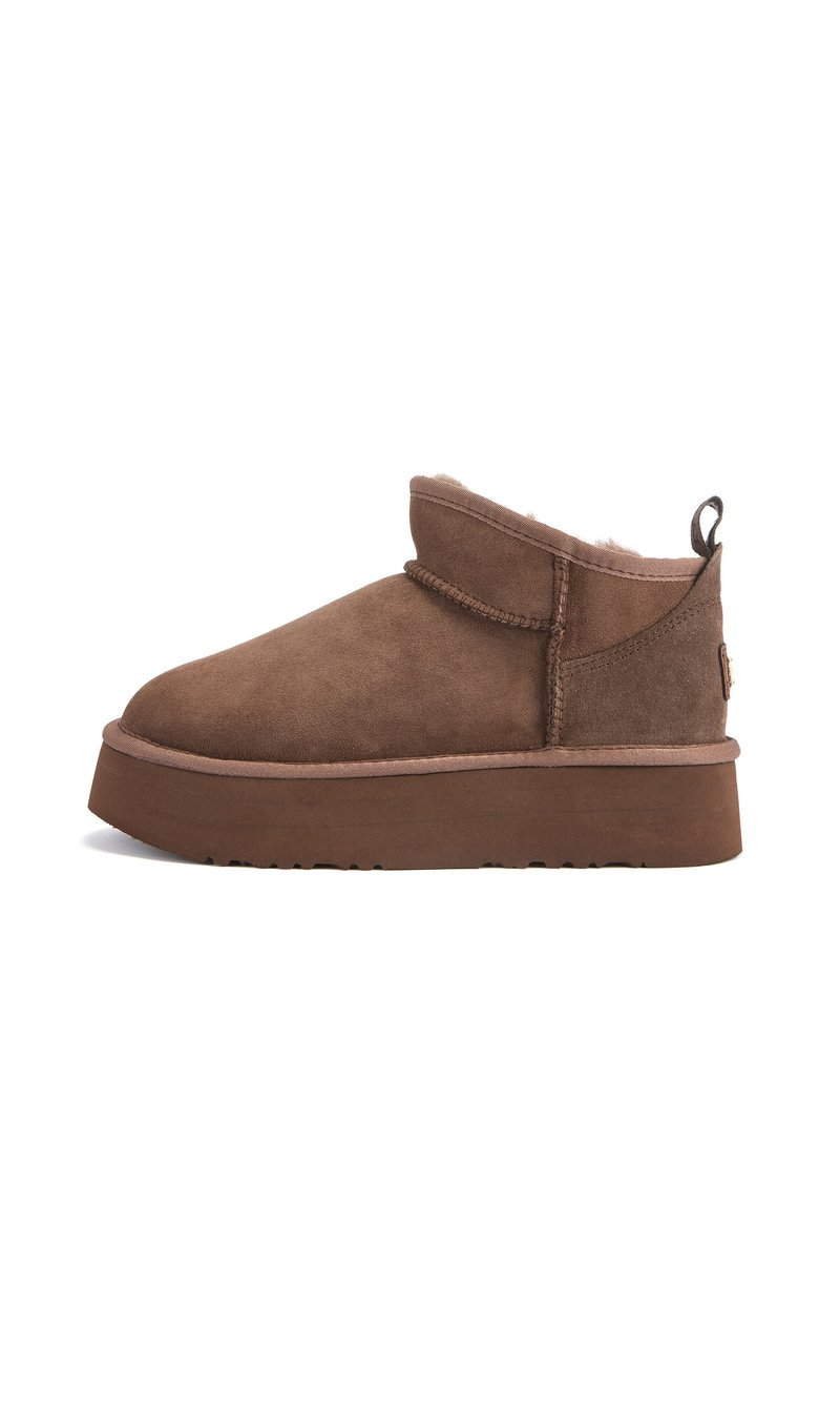 Botas de ante marrón hasta el tobillo con punta redonda, suela plana y forro de piel de oveja. Presentan detalles cosidos y una pestaña para el talón.