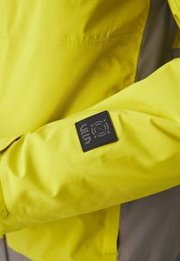 Chaqueta impermeable amarilla con textura suave, presenta un parche de logo negro en la manga, destacando un diseño moderno y alta visibilidad.