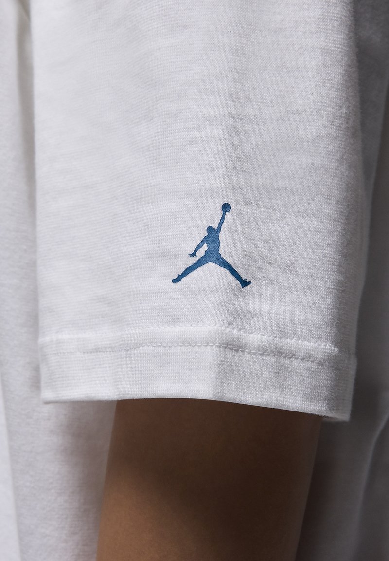 Maglietta bianca a maniche corte realizzata in tessuto texturizzato con il logo Jumpman blu stampato sulla manica sinistra. Presenta una vestibilità standard e orlo cucito.