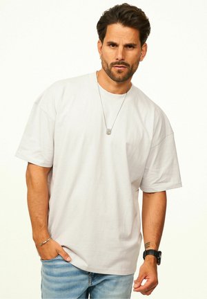 Mann mit Bart, der ein schlichtes weißes, übergroßes T-Shirt, blaue Jeans, eine Halskette, ein Armband und eine schwarze Armbanduhr vor einem schlichten Hintergrund trägt.