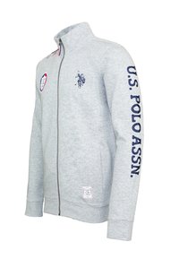 Grå zip-up sweatshirt i mjukt tyg. Har en blå U.S. Polo Assn. logotyp på ärmen och ett märke på bröstet. Inkluderar två sidfickor.
