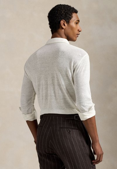 Ανοιχτό μπεζ πλεκτό πόλο μπλουζάκι με γιακά, ψηλοτά ανασηκωμένα μανίκια και άνετη εφαρμογή, σε συνδυασμό με σκούροριγέ παντελόνι με πίσω τσέπες.