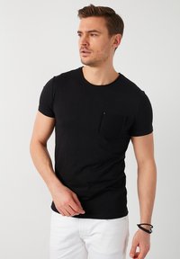 T-shirt nera a maniche corte in cotone con scollatura rotonda e una tasca a toppa testurizzata sul lato sinistro del petto. Il modello la abbina a jeans bianchi.