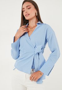 Donna che indossa una blusa avvolgente a maniche lunghe a righe blu e bianche con cintura in vita, abbinata a pantaloni bianchi e gioielli dorati.