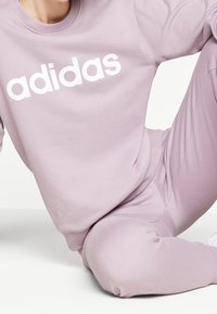 Světle fialová mikina Adidas s bílým logem, kulatým výstřihem a měkkou, strukturovanou tkaninou, v kombinaci s odpovídajícími kalhotami.