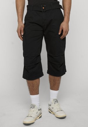 Hombre con pantalones cortos cargo negros hasta la rodilla, calcetines blancos hasta el tobillo y zapatillas deportivas blancas, de pie frente a un fondo liso.