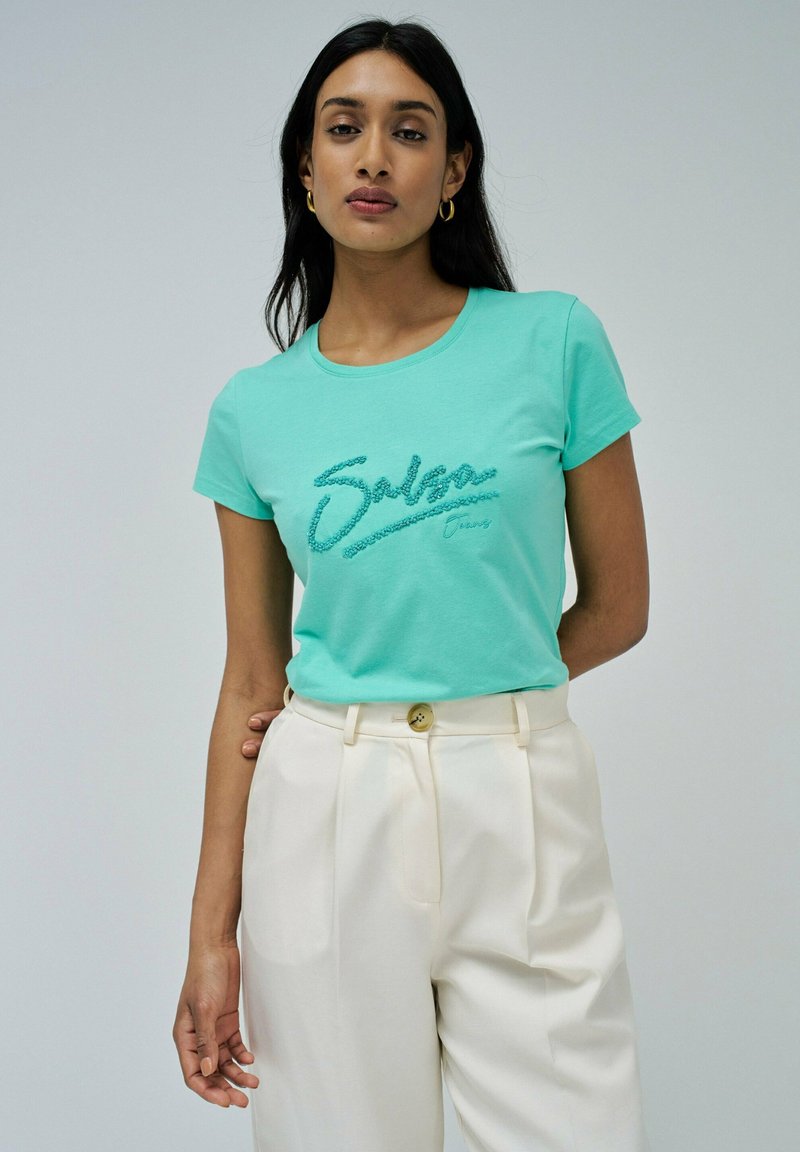 Camiseta de manga corta verde menta con letras texturizadas "Salsa"; combinada con pantalones crema y un botón dorado en la cintura.