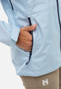 Hellblaue Jacke mit einer Reißverschlusstasche, dunklen Reißverschlüssen und verstellbarem Kordelzug am Saum. Der Stoff sieht glatt und leicht aus.