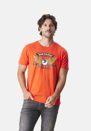 T-shirt en coton orange à manches courtes avec un motif imprimé de ailes et d'un œil. Jeans gris visibles à la taille.