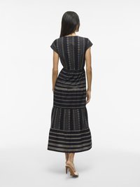 A woman stands with her back to the camera, wearing a long, patterned dress with cap sleeves and heeled sandals.