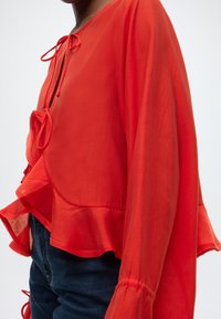 Κόκκινη, ελαφριά μπλούζα με μακριά, φαρδιά μανίκια, λαιμόκοψη με κορδόνι και μπορντούρα με βολάν. Το ύφασμα είναι διαφανές με απαλή υφή.