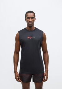 Črna majica brez rokavov z mehko teksturo in logotipom "HUGO" v rdeče-sivem prelivu. Kombinirana s temnimi kratkimi hlačami.