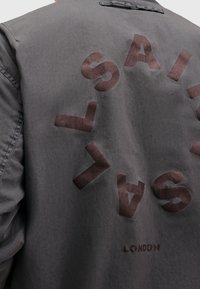 Parte trasera de una chaqueta gris con letras circulares en marrón oscuro que dicen "LASAK" y un pequeño texto "LONDON" debajo, todo sobre la tela.