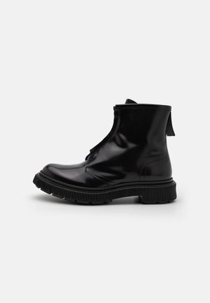 Botas de tobillo negras brillantes con una suela texturizada, punta redondeada y detalle de cremallera. Cuenta con un tirador en la parte trasera para facilitar su uso.