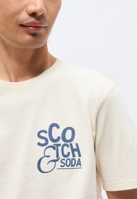 Νέος άντρας που φοράει ένα κρεμ t-shirt με μπλε λογότυπο "Scotch & Soda" στο στήθος, μπροστά σε απλό φωτεινό φόντο.