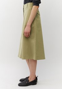 Persoon in een olijfgroene midi-rokken, zwarte loafers en een zwart top, staande zijwaarts tegen een eenvoudige witte achtergrond.