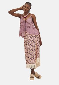 Blusa púrpura con volantes y lazos en el frente, combinada con una falda con estampado de flores y flecos en el dobladillo. Sandalias de plataforma beige.
