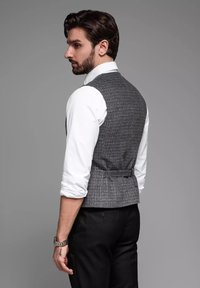 Grijze geblokte gilet met een aansluitende pasvorm, gecombineerd met een witte blouse. Bevat een taille-aanpassingsriem en een gladde textuur. Zwarte broek zichtbaar.